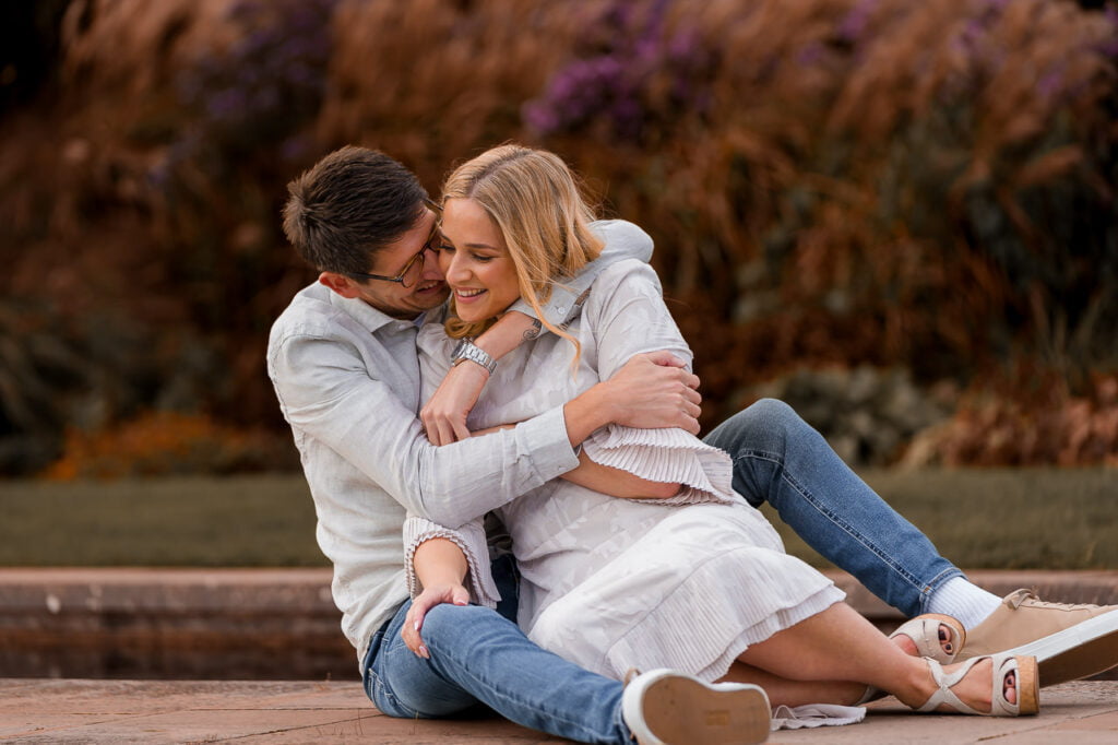 engaged couple playing around and cuddling together while sitting on the payment way through the water in the gardens of their summer engagement
