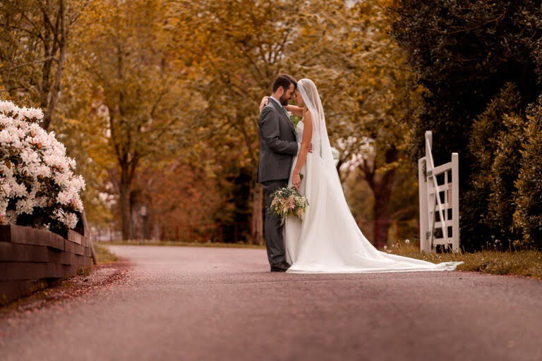 A bride and groom stand on a tree-lined path surrounded by autumn foliage at what was Wasing-Park-Spring wedding in Berkshire. The bride, in a white dress with a long veil, holds a bouquet as the groom, in a gray suit, embraces her under the soft golden light.
