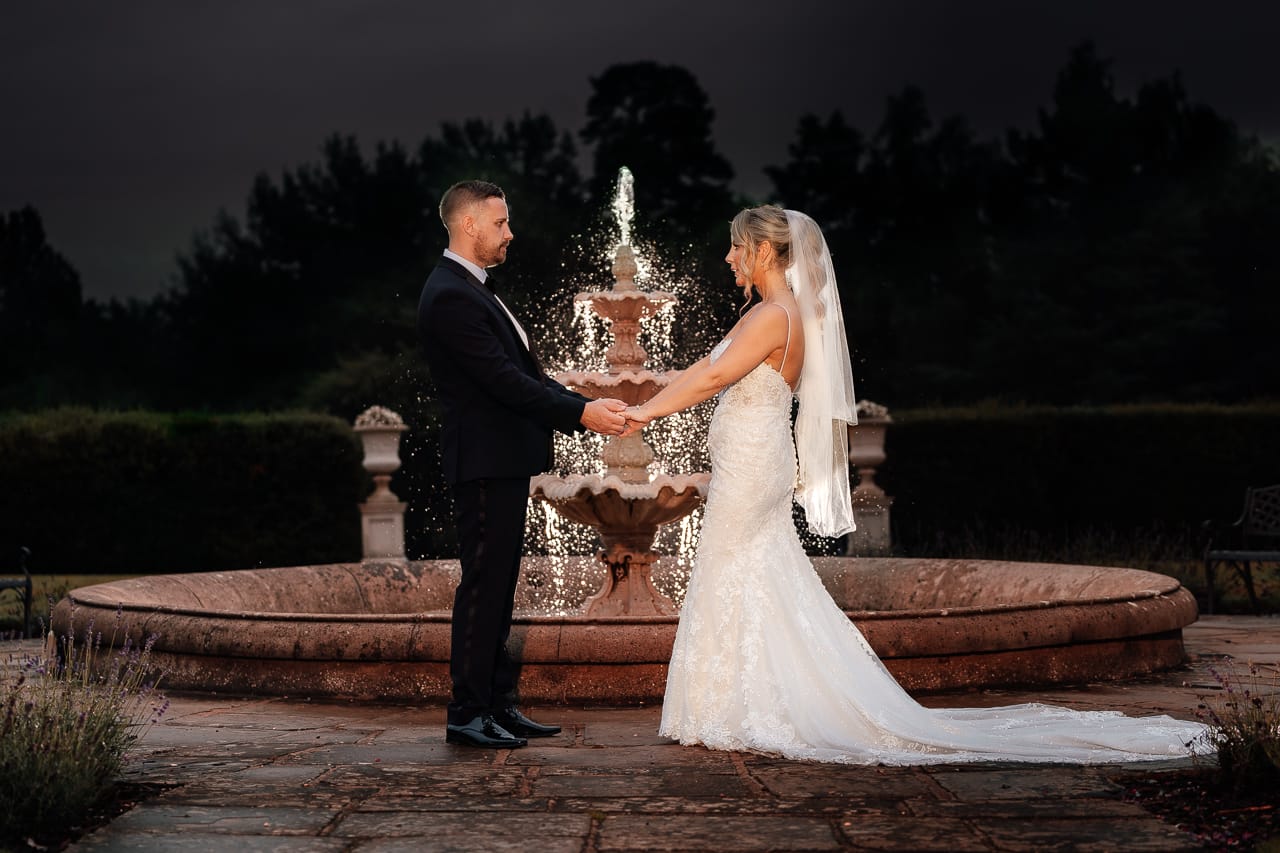 A bride and groom stand facing each other, holding hands in front of an illuminated fountain at night, with trees and dark sky in the background at their elegant Easthampstead Park summer wedding.
