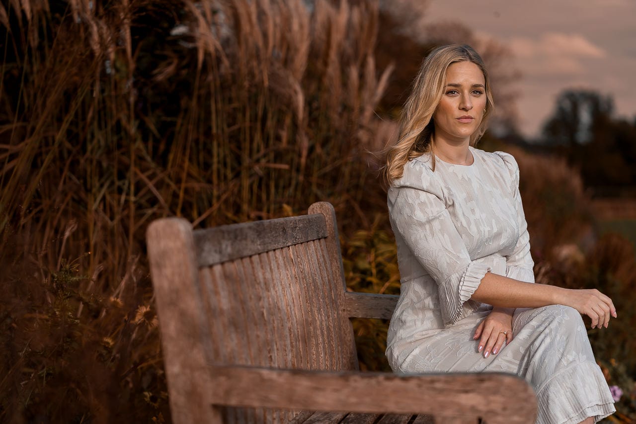 woman sitting on a wooden bench with a white jumpsuit and arms folded over her legs, posing for her summer engagement session