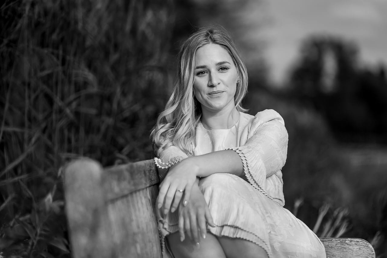 black and white photo of a woman sitting on a wooden bench with a white jumpsuit and arms folded over her legs, posing for her summer engagement session