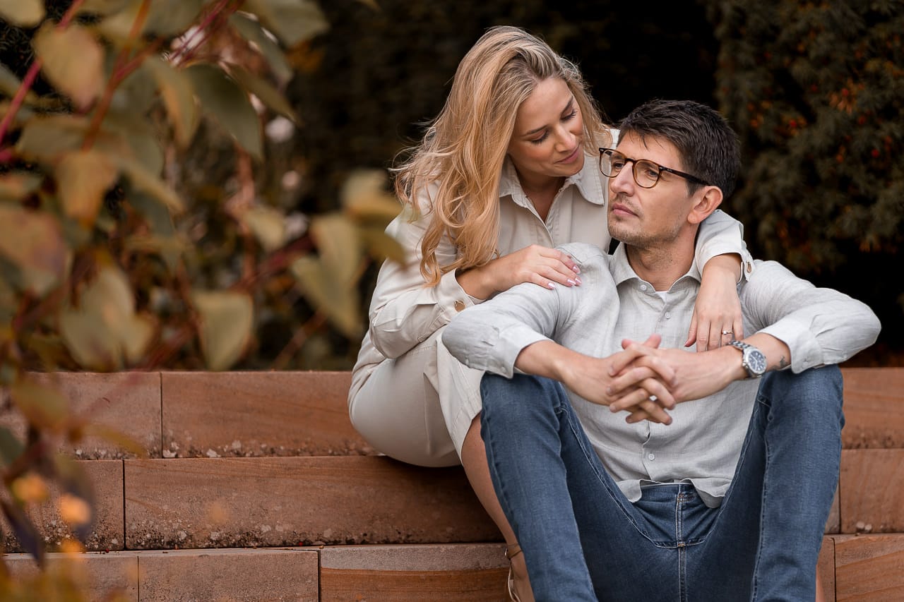 summer engagement of couple sitting on steps with woman behind her and arms laying over his shoulders